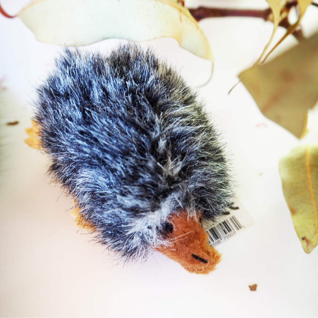 A gray echidna finger puppet nestled in a natural-looking grassy nest, with leaves and a wooden surface in the background.