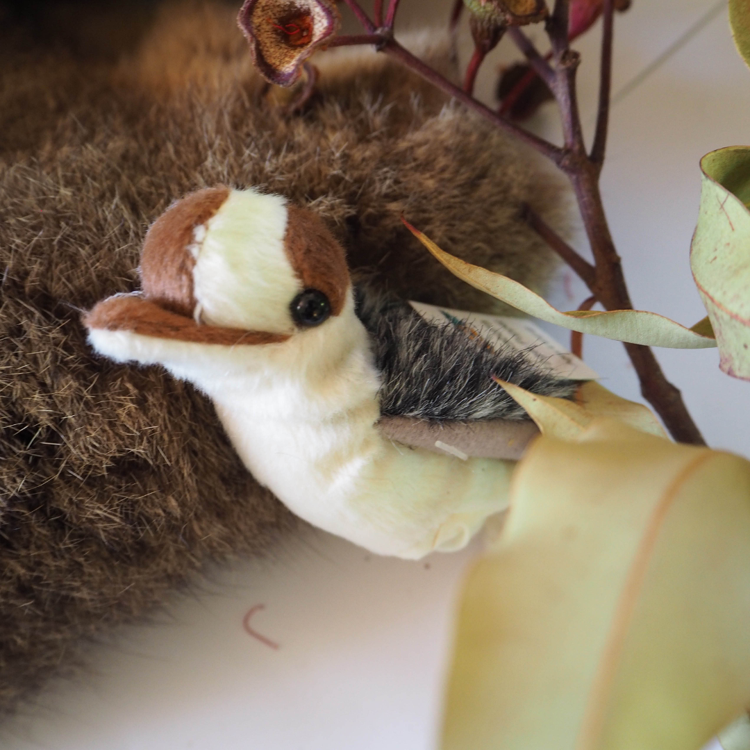 A plush finger puppet designed to look like a kookaburra bird, placed on a wooden surface with a background of potted plants and red flowers.
