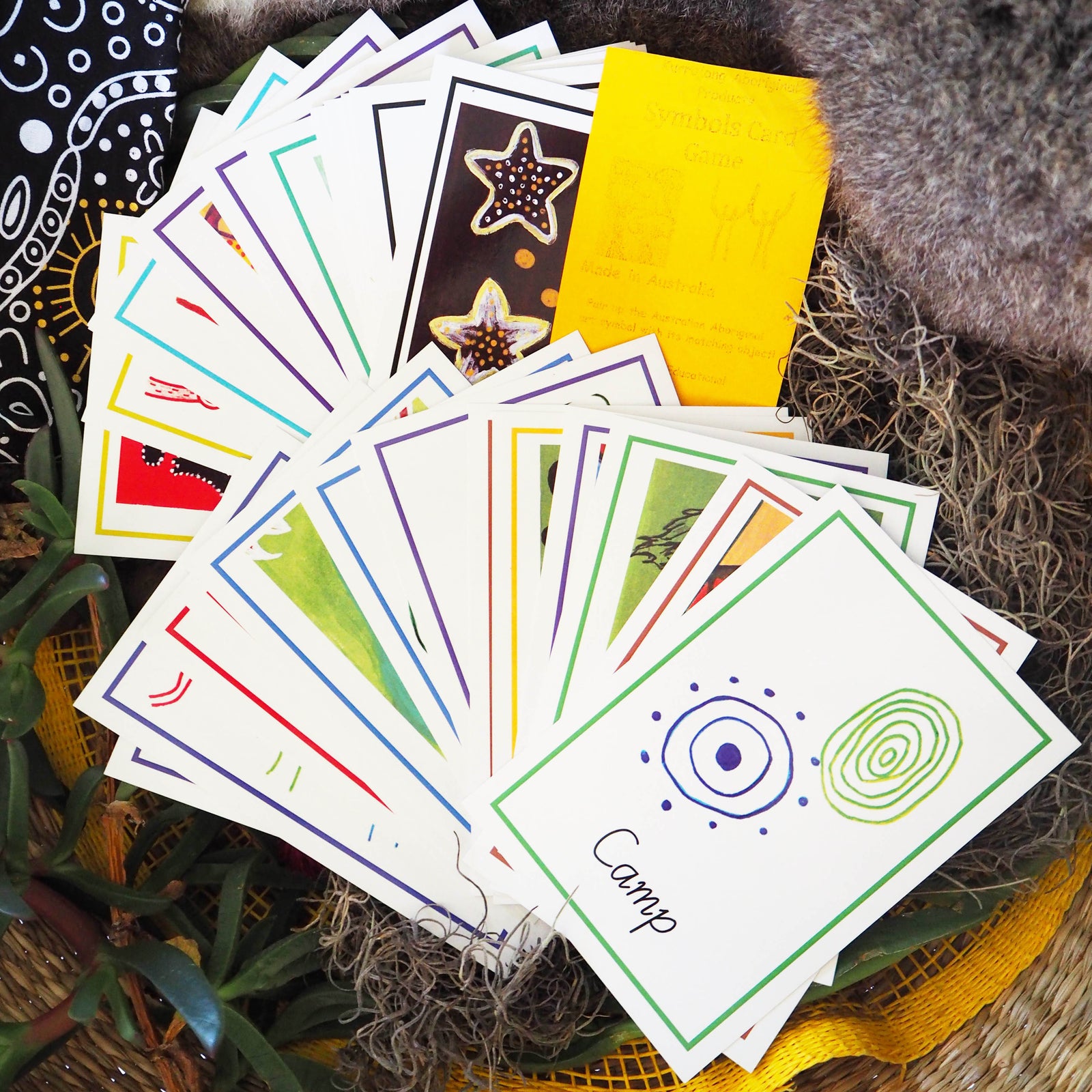 A collection of colorful Aboriginal symbol cards spread out, with some cards showing geometric patterns and others with illustrations, placed on a textured surface.